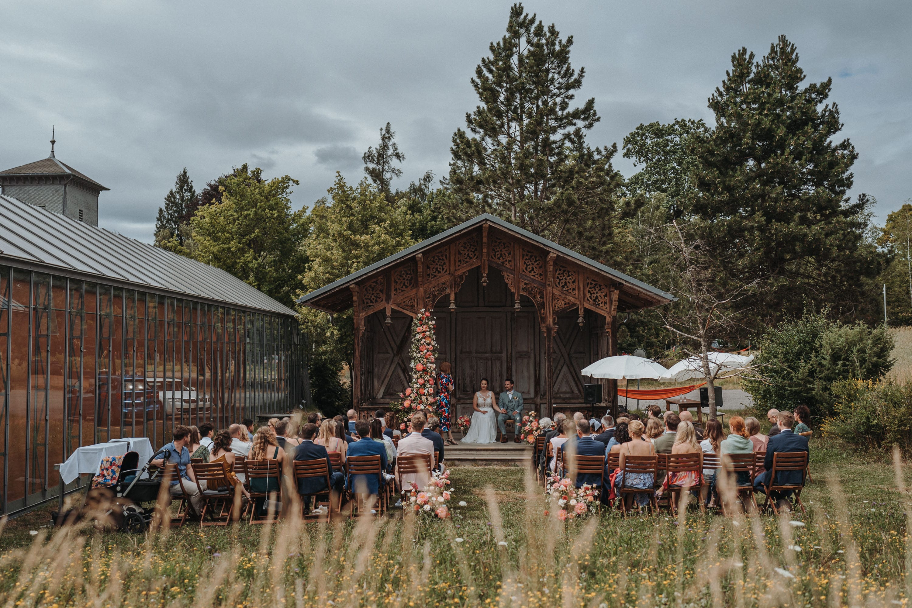 Freie Trauung vor dem geschnitzten Holzpavillon, vorne hohes Gras