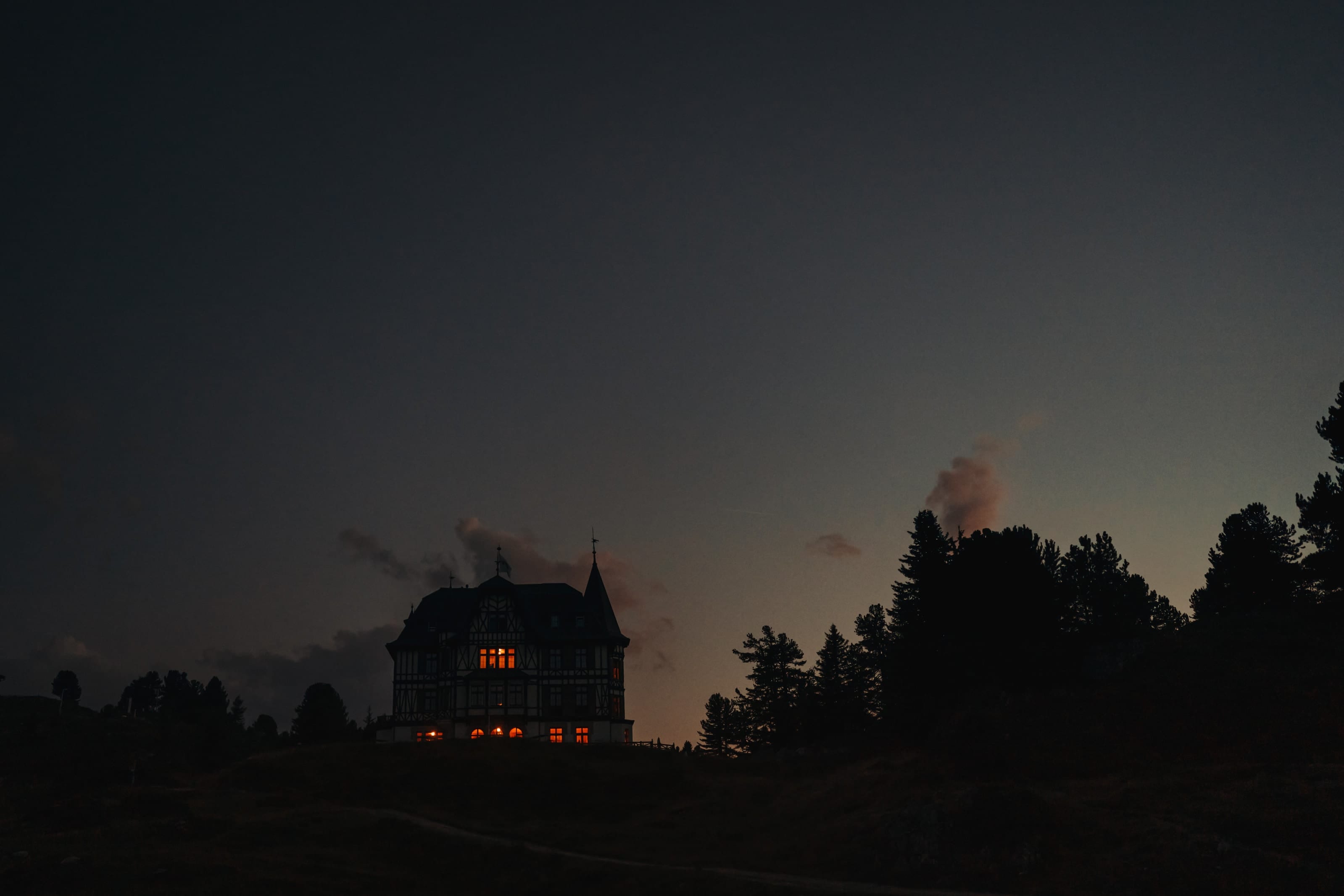 Ein historisches Haus in der Dämmerung, klein am unteren Bildrand, einige Fenster leuchten warm unter einem grossen dunklen Himmel